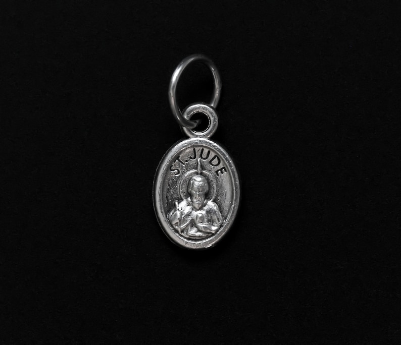 May include: Silver charm with a depiction of Saint Jude Thaddeus, patron saint of hopeless causes. The charm is oval shaped and has the words "St. Jude" inscribed around the image.