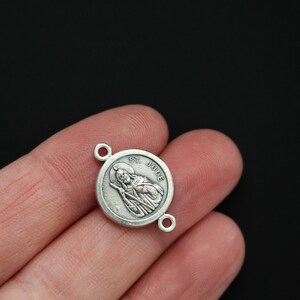 May include: Silver-toned St. Jude Thaddeus charm with a round shape and a detailed image of the saint. The charm has two jump rings for attaching to a necklace or bracelet.
