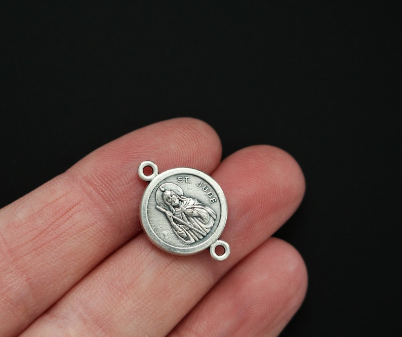 May include: Silver-toned St. Jude Thaddeus charm with a round shape and a detailed image of the saint. The charm has two jump rings for attaching to a necklace or bracelet.