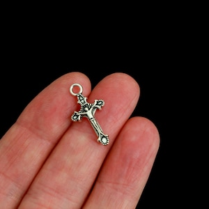 May include: A small silver-toned metal crucifix charm featuring an intricate design with a central figure of Jesus. The cross has decorative flared ends and a circular jump ring at the top for attachment, showcasing a polished and oxidized finish.