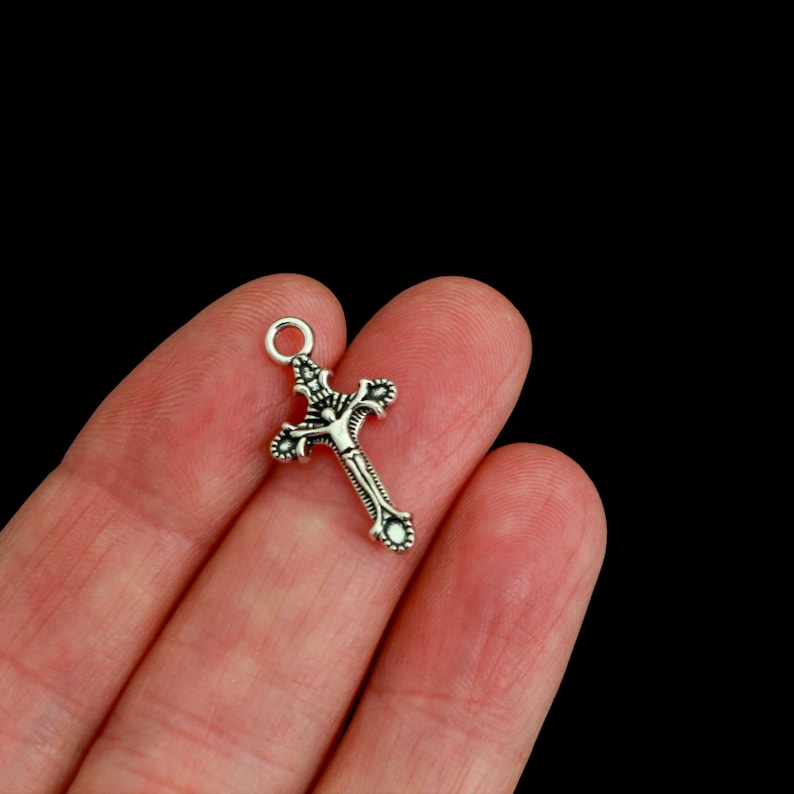 May include: A small silver-toned metal crucifix charm featuring an intricate design with a central figure of Jesus. The cross has decorative flared ends and a circular jump ring at the top for attachment, showcasing a polished and oxidized finish.