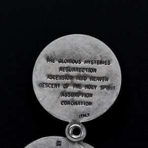May include: A silver religious pendant with two circular sections. The top section has the text "THE GLORIOUS MYSTERIES" and other religious terms. The word "ITALY" is at the bottom. The pendant has a small hole for attachment.