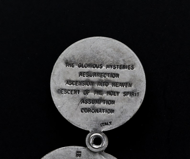May include: A silver religious pendant with two circular sections. The top section has the text "THE GLORIOUS MYSTERIES" and other religious terms. The word "ITALY" is at the bottom. The pendant has a small hole for attachment.