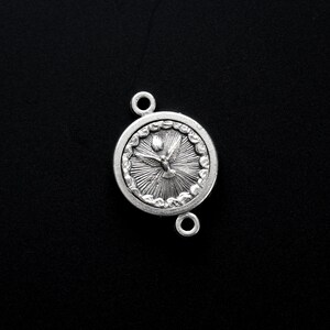 May include: A silver-colored round pendant with a dove and sunburst design. The pendant has two loops for attachment and is set against a black background. The dove is depicted with outstretched wings.