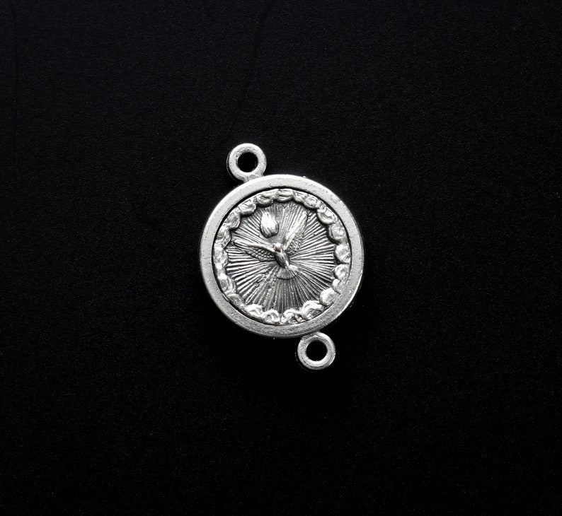 May include: A silver-colored round pendant with a dove and sunburst design. The pendant has two loops for attachment and is set against a black background. The dove is depicted with outstretched wings.