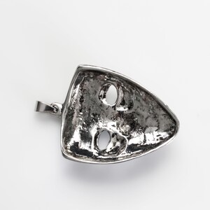 May include: Silver pendant in the shape of a face mask with two eye holes. The pendant has a textured surface and a loop for attaching a chain.