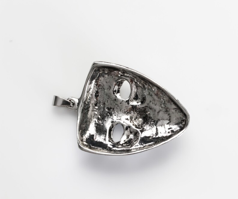 May include: Silver pendant in the shape of a face mask with two eye holes. The pendant has a textured surface and a loop for attaching a chain.
