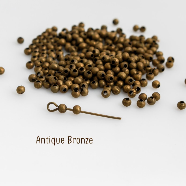 May include: A collection of small, round, antique bronze metal beads scattered across a surface, accompanied by a single metal headpin featuring two attached beads and a loop at one end. The text Antique Bronze is printed below.
