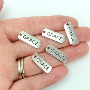 May include: Five silver metal tags with the word "GRACE" engraved on them. The tags have a hole at the top for attaching to a necklace or bracelet.