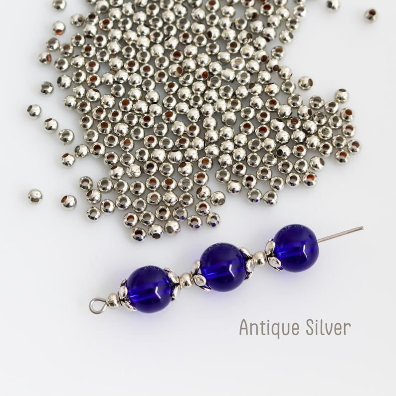 May include: A collection of small, round, metallic silver-colored spacer beads scattered across the surface, alongside a single metal wire strand featuring three deep blue spherical beads separated by ornate antique silver-colored bead caps.