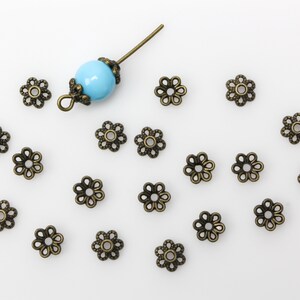 Puede incluir: Una colecci&oacute;n de 18 tapas de cuentas de bronce antiguo en forma de flor con una sola cuenta azul turquesa y un pasador de metal.