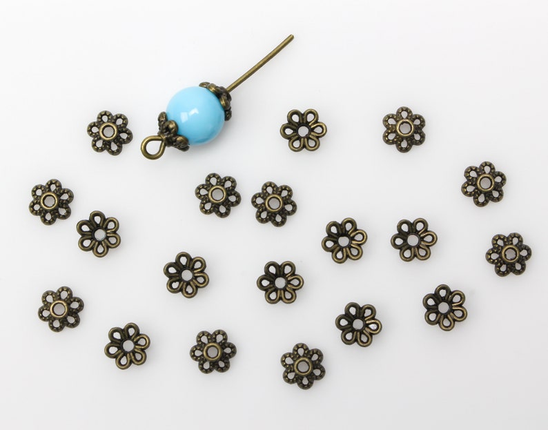 Puede incluir: Una colecci&oacute;n de 18 tapas de cuentas de bronce antiguo en forma de flor con una sola cuenta azul turquesa y un pasador de metal.