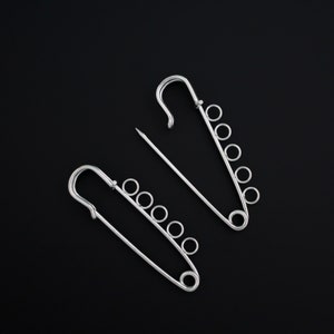 May include: Two silver-colored safety pins with attached small rings, set against a black background. One pin is open, with the needle separate, while the other is closed. The pins are designed with a decorative element of small rings.