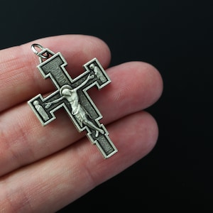 Op de afbeelding: Een zilverkleurige kruis hanger met een gedetailleerde afbeelding van Jezus Christus aan het kruis. Het kruis heeft ingewikkelde ontwerpen en een lus aan de bovenkant om op te hangen.