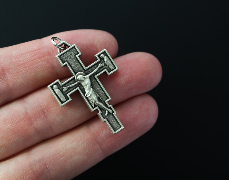 Op de afbeelding: Een zilverkleurige kruis hanger met een gedetailleerde afbeelding van Jezus Christus aan het kruis. Het kruis heeft ingewikkelde ontwerpen en een lus aan de bovenkant om op te hangen.