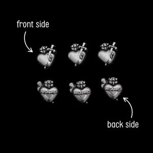 May include: Six silver-toned heart-shaped charms with a crown and cross design. The charms are arranged on a black background, with arrows indicating the front and back sides. The charms have a detailed, textured surface.