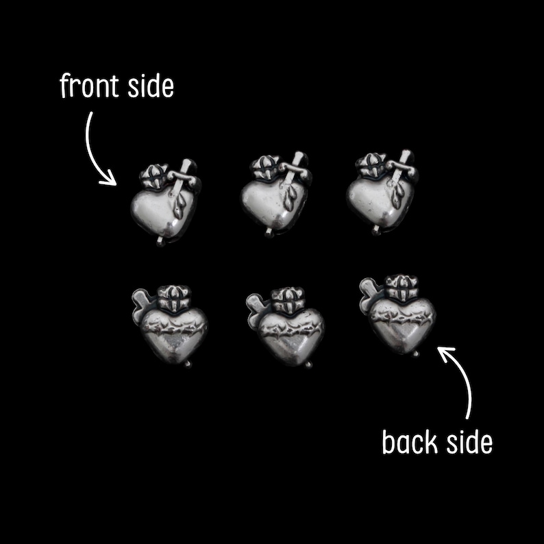 May include: Six silver-toned heart-shaped charms with a crown and cross design. The charms are arranged on a black background, with arrows indicating the front and back sides. The charms have a detailed, textured surface.