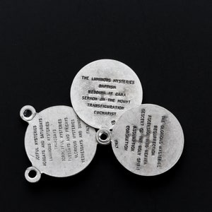 May include: Three silver religious medallions with text on a black background. The medallions are arranged in a circular pattern. Each lists religious mysteries and events, including "Baptism" and "Resurrection". The word "Italy" is also visible.