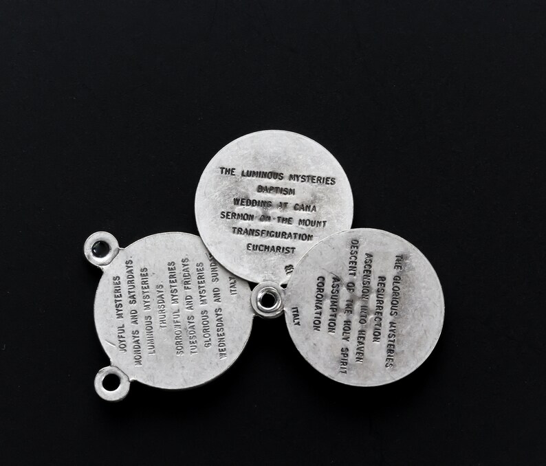 May include: Three silver religious medallions with text on a black background. The medallions are arranged in a circular pattern. Each lists religious mysteries and events, including "Baptism" and "Resurrection". The word "Italy" is also visible.