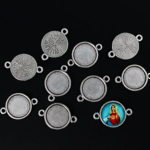 May include: Ten silver-toned metal round pendant trays with a textured surface. The trays have two loops on each side for attaching to a chain or cord. One tray features a colourful image of a woman with a heart.