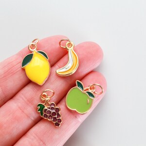 Pu&ograve; includere: Quattro ciondoli dorati che raffigurano diversi frutti: un limone giallo, una banana gialla, una mela verde e un grappolo d'uva viola.