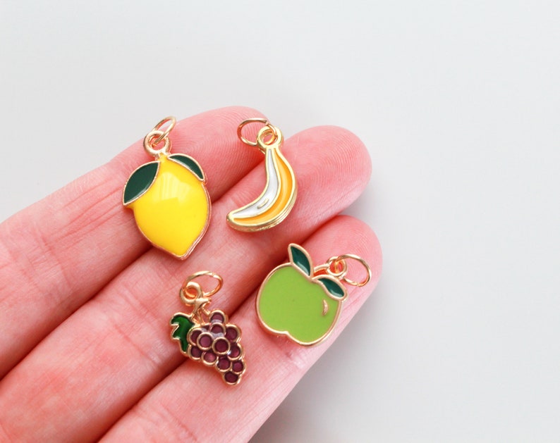 Pu&ograve; includere: Quattro ciondoli dorati che raffigurano diversi frutti: un limone giallo, una banana gialla, una mela verde e un grappolo d'uva viola.