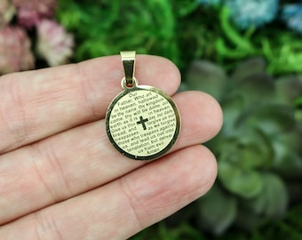 May include: Gold-toned pendant with a round, engraved disc featuring the Lord's Prayer. The pendant is held in a hand against a green background.