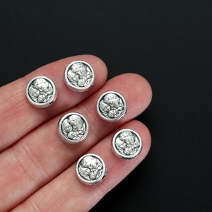 May include: Six silver-toned round beads with a raised design of a face. The beads are on a hand, and the background is black.