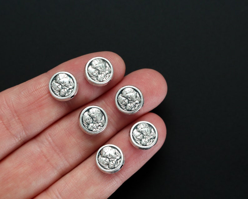 May include: Six silver-toned round beads with a raised design of a face. The beads are on a hand, and the background is black.