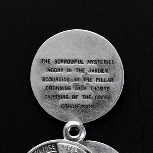 May include: A silver religious pendant with two circular sections. The upper disc has the text "THE SORROWFUL MYSTERIES" and other religious phrases. The pendant is set against a black backdrop.