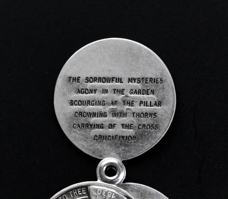 May include: A silver religious pendant with two circular sections. The upper disc has the text "THE SORROWFUL MYSTERIES" and other religious phrases. The pendant is set against a black backdrop.