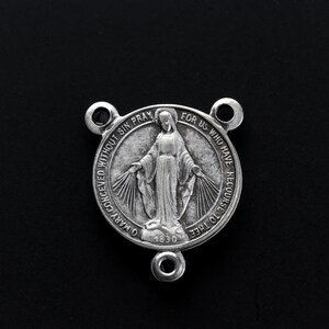 May include: A silver religious medal featuring a woman with outstretched arms, encircled by text. The medal has three attachment loops and the year 1830 is inscribed on it.