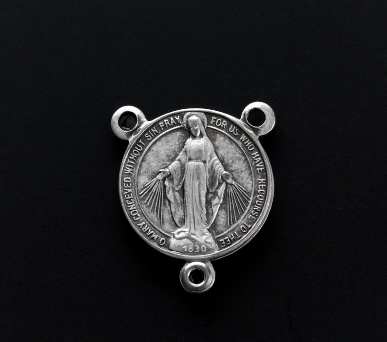 May include: A silver religious medal featuring a woman with outstretched arms, encircled by text. The medal has three attachment loops and the year 1830 is inscribed on it.