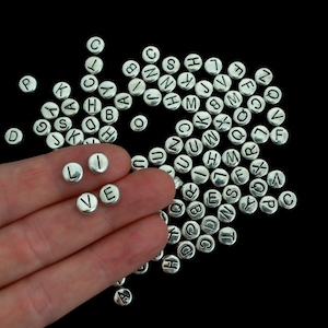 May include: Silver-colored, round alphabet beads scattered on a black surface. The beads are engraved with letters from A to Z. Some beads spell the word "LOVE". Ideal for jewelry making and crafting.