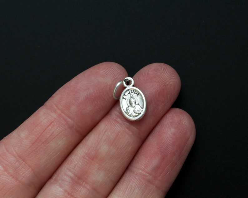 May include: Silver charm with a depiction of Saint Jude Thaddeus, a Catholic saint, and the words "St. Jude" inscribed around the image.