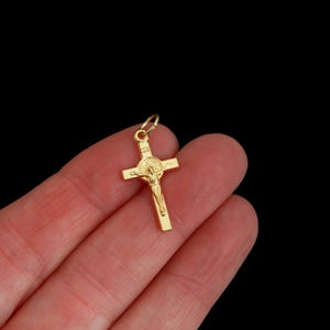 Op de afbeelding: Gouden kruisbeeldhanger met een gedetailleerde afbeelding van Jezus Christus aan het kruis. De hanger heeft een kleine lus aan de bovenkant voor een ketting. Zwarte achtergrond.