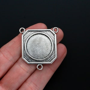 May include: Silver-toned metal pendant with a round, blank space in the center. The pendant has a decorative, octagonal shape with a raised edge around the blank space. There are three loops on the pendant for attaching a chain or cord.
