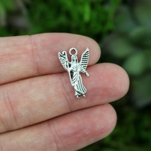 May include: Silver charm featuring a detailed angel with wings outstretched. The angel is standing with its arms crossed and has a halo above its head.