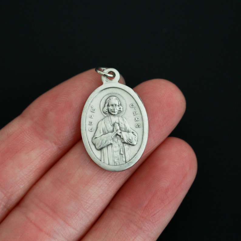 May include: Silver oval pendant with a raised image of a man in a robe with his hands clasped in prayer. The text "CUR&Eacute; D'ARS" is inscribed around the edge of the pendant.