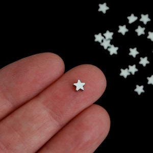 May include: A small, silver-colored star charm rests on a fingertip, with several more scattered against a black background. The star has five points and a smooth, reflective surface. This image showcases a collection of star-shaped charms.