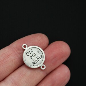 May include: Silver charm with the words "Ora Pro Nobis" and "Italy" engraved on it. The charm has two loops for attaching to a bracelet or necklace.