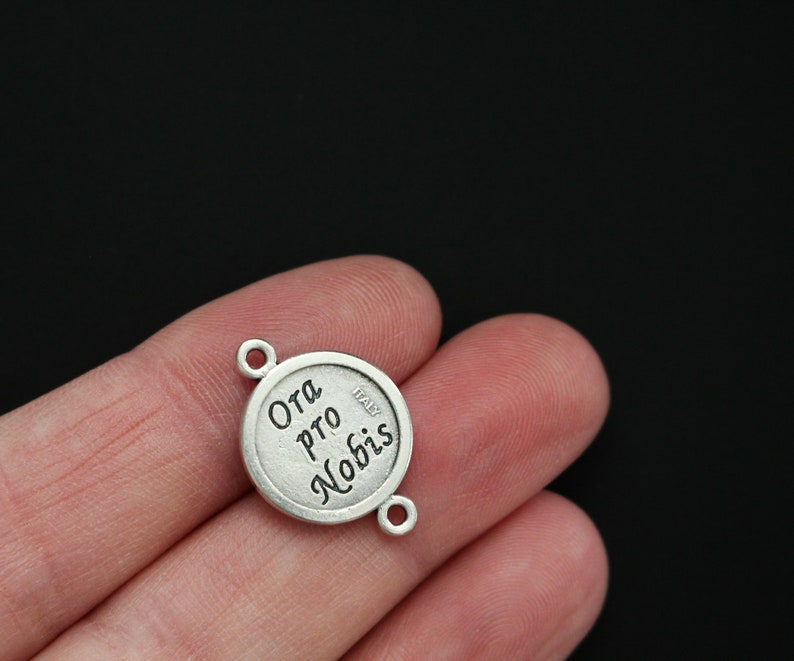May include: Silver charm with the words "Ora Pro Nobis" and "Italy" engraved on it. The charm has two loops for attaching to a bracelet or necklace.