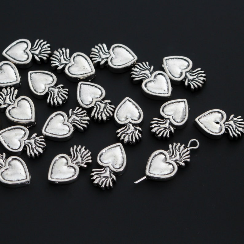 May include: A collection of silver-toned metal heart charms with a flame design. The charms are scattered on a black surface.