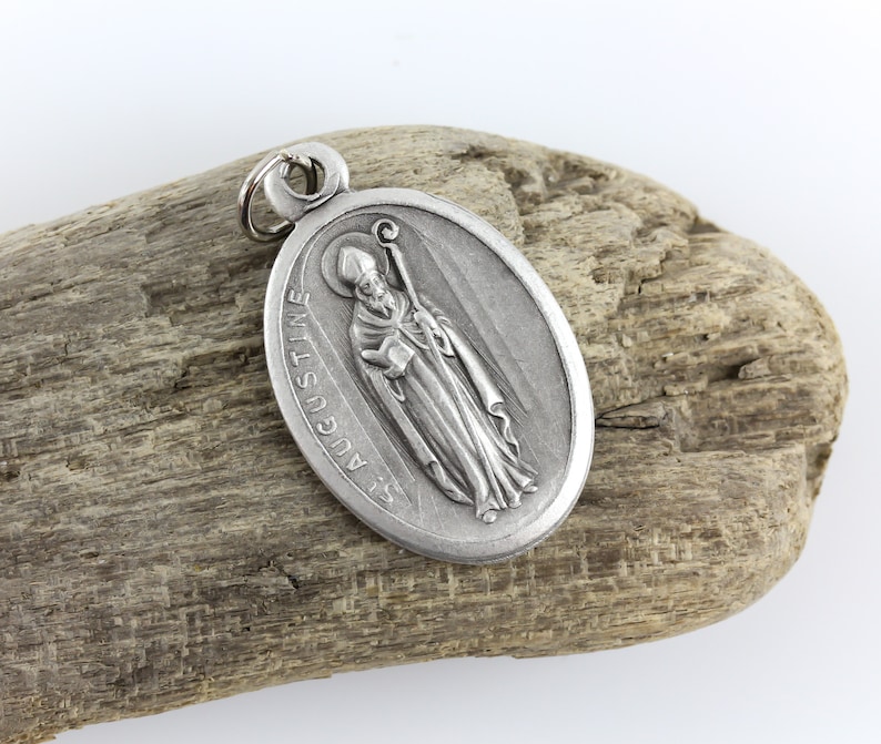 May include: Silver oval pendant with a depiction of Saint Augustine, a Catholic saint, holding a book and a staff. The pendant has the words "St. Augustine" inscribed around the edge.