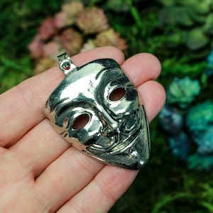 Op de afbeelding: Een zilveren metalen hanger in de vorm van een masker met een ondeugende grijns. Het masker heeft twee ooggaten en een brede, tanden grijns.