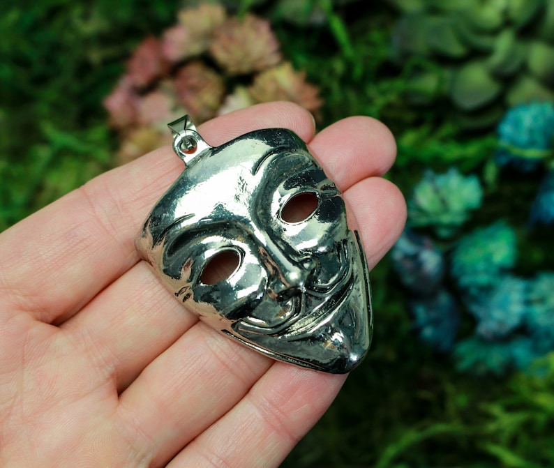May include: A silver metal pendant in the shape of a mask with a mischievous grin. The mask has two eye holes and a wide, toothy smile.