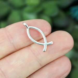 Puede incluir: Un colgante plateado con forma del símbolo Ichthys, también conocido como pez de Jesús. El colgante es un contorno de pez estilizado con un lazo en la parte superior para una cadena. El fondo está borroso con tonos verdes y azules.