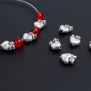 May include: Silver heart-shaped beads with a small crown design are scattered on a black surface. A strand of red glass beads and silver heart beads is visible. The beads are likely for jewelry making.