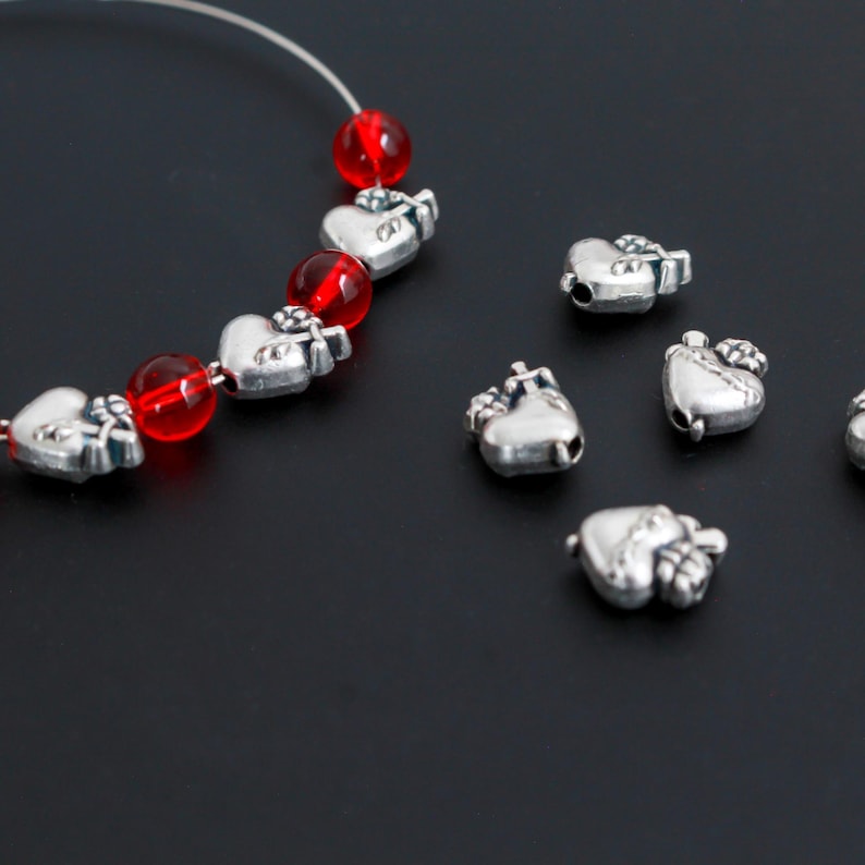 May include: Silver heart-shaped beads with a small crown design are scattered on a black surface. A strand of red glass beads and silver heart beads is visible. The beads are likely for jewelry making.