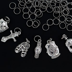 May include: A collection of silver-toned charms featuring various religious symbols, including a cross, a shield, a helmet, and a vest. The charms are arranged on a black background with a pile of silver jump rings.